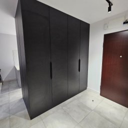 Nina Meble - Czarna, matowa zabudowa szafy wnękowej z długimi, pionowymi uchwytami, dopasowana do wysokości pomieszczenia, na tle jasnej podłogi z płytek i białych ścian.