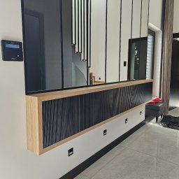 Nina Meble - Nowoczesna zabudowa ściany z lamelami w kolorze czarnym, obudowana jasnym drewnem, z przeszklonym panelem górnym, odbijającym schody i fragment wnętrza.