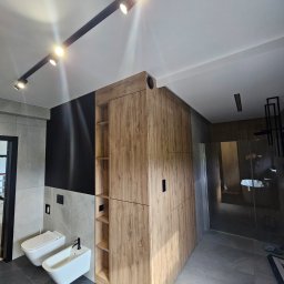 Nina Meble - Nowoczesna łazienka z drewnianą szafą wnękową, podwieszaną toaletą i bidetem, oświetlenie szynowe punktowe.