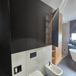 Nina Meble - Nowoczesna łazienka z czarnymi szafkami wiszącymi, otwartą szafką z drewnianym fornirem, białą toaletą i bidetem na tle szarych płytek.