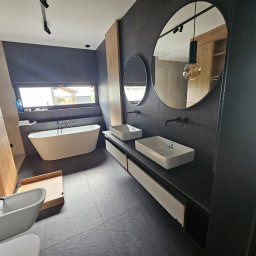 Nina Meble - Nowoczesna łazienka z czarnymi płytkami, dwiema umywalkami nablatowymi, okrągłymi lustrami, wolnostojącą wanną przy oknie i drewnianą zabudową szaf.