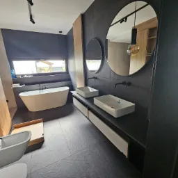 Nowoczesna łazienka z czarnymi płytkami, dwiema umywalkami nablatowymi, okrągłymi lustrami, wolnostojącą wanną przy oknie i drewnianą zabudową szaf.