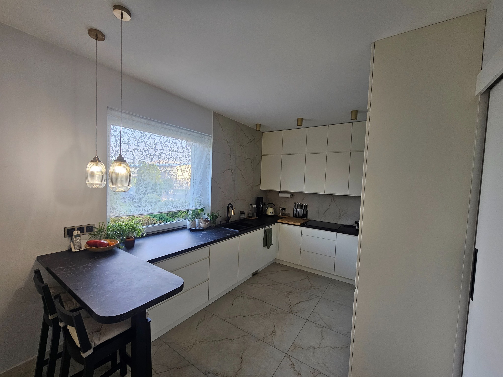 Jasna kuchnia z czarnym blatem i marmurowymi płytkami na ścianie, oświetlona wiszącymi lampami z żarówkami dekoracyjnymi, z dodatkowym blatem barowym i krzesłami.