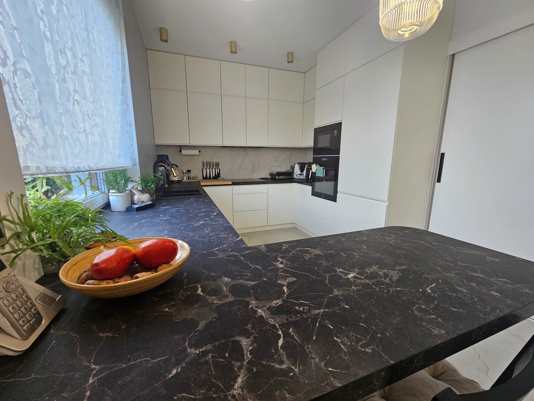 Nowoczesna, biała kuchnia z czarnym blatem marmurkowym, owocami w misie i widokiem z okna.