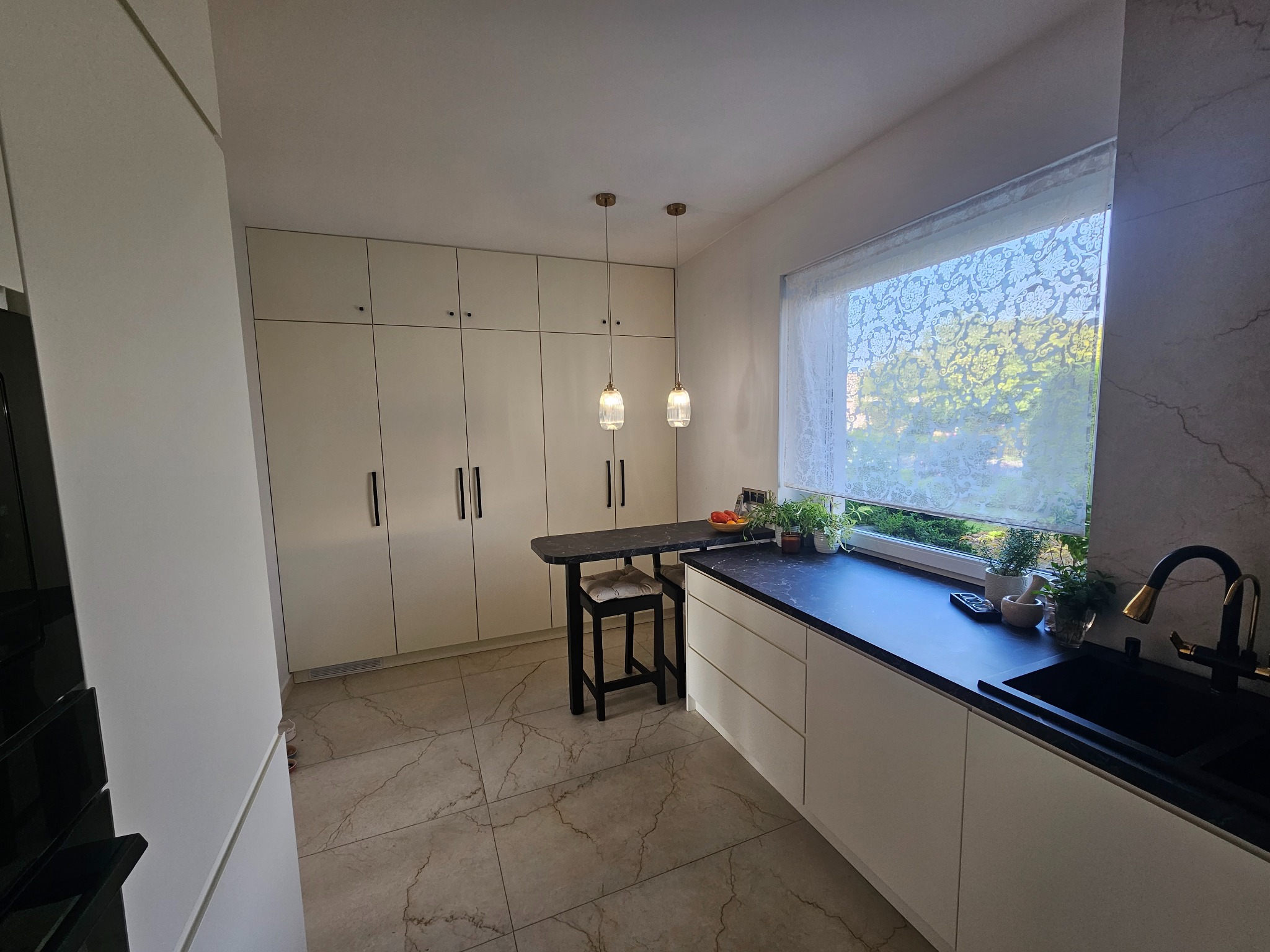 Nowoczesna kuchnia z białymi frontami szafek, czarnym blatem, marmurową ścianą nad zlewem, wiszącymi lampami ze złotymi elementami i stołkiem barowym.