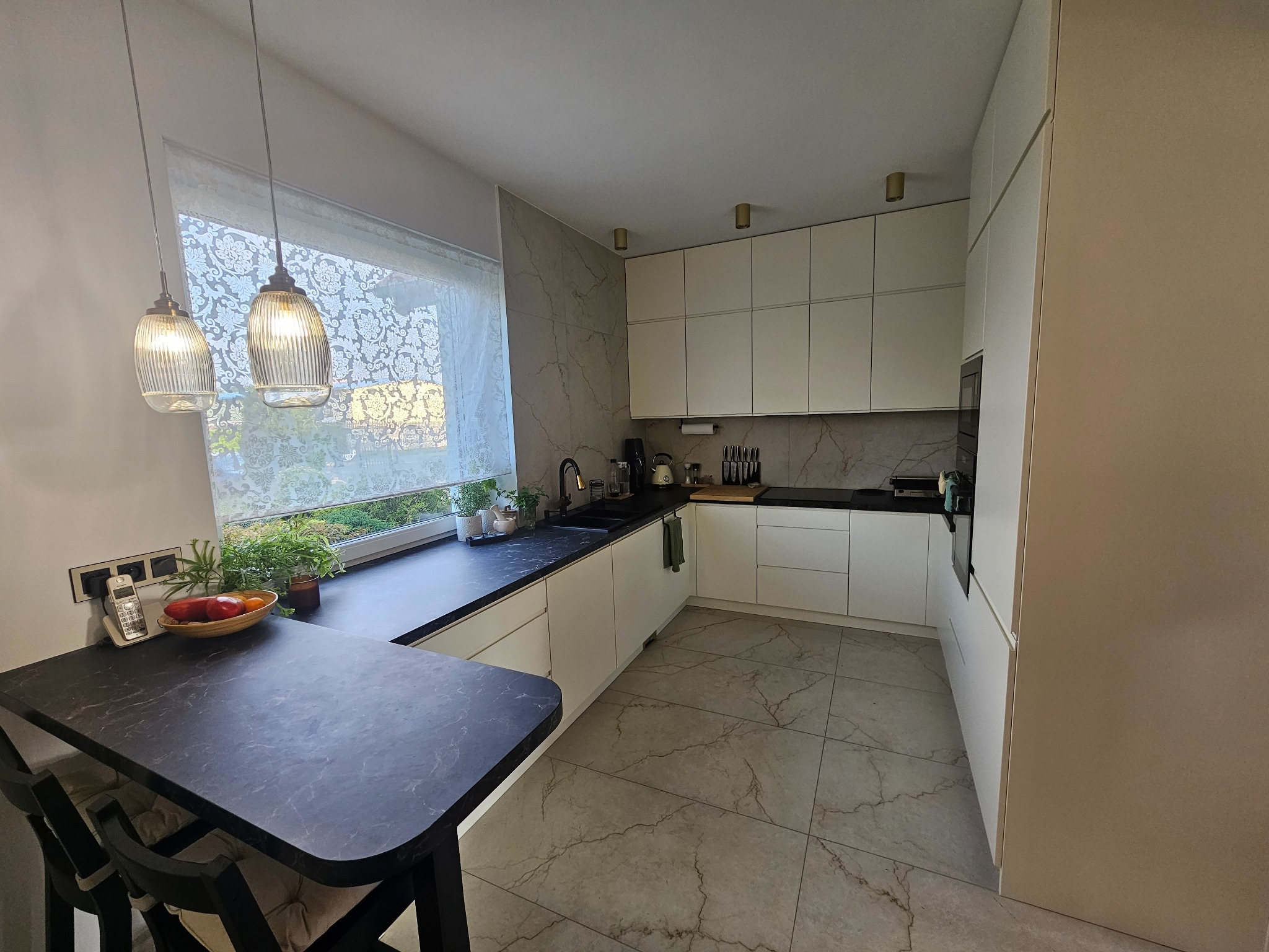 Nowoczesna kuchnia z białymi szafkami, czarnym blatem, marmurową ścianą i podłogą, oświetlona wiszącymi lampami nad stołem z owocami.