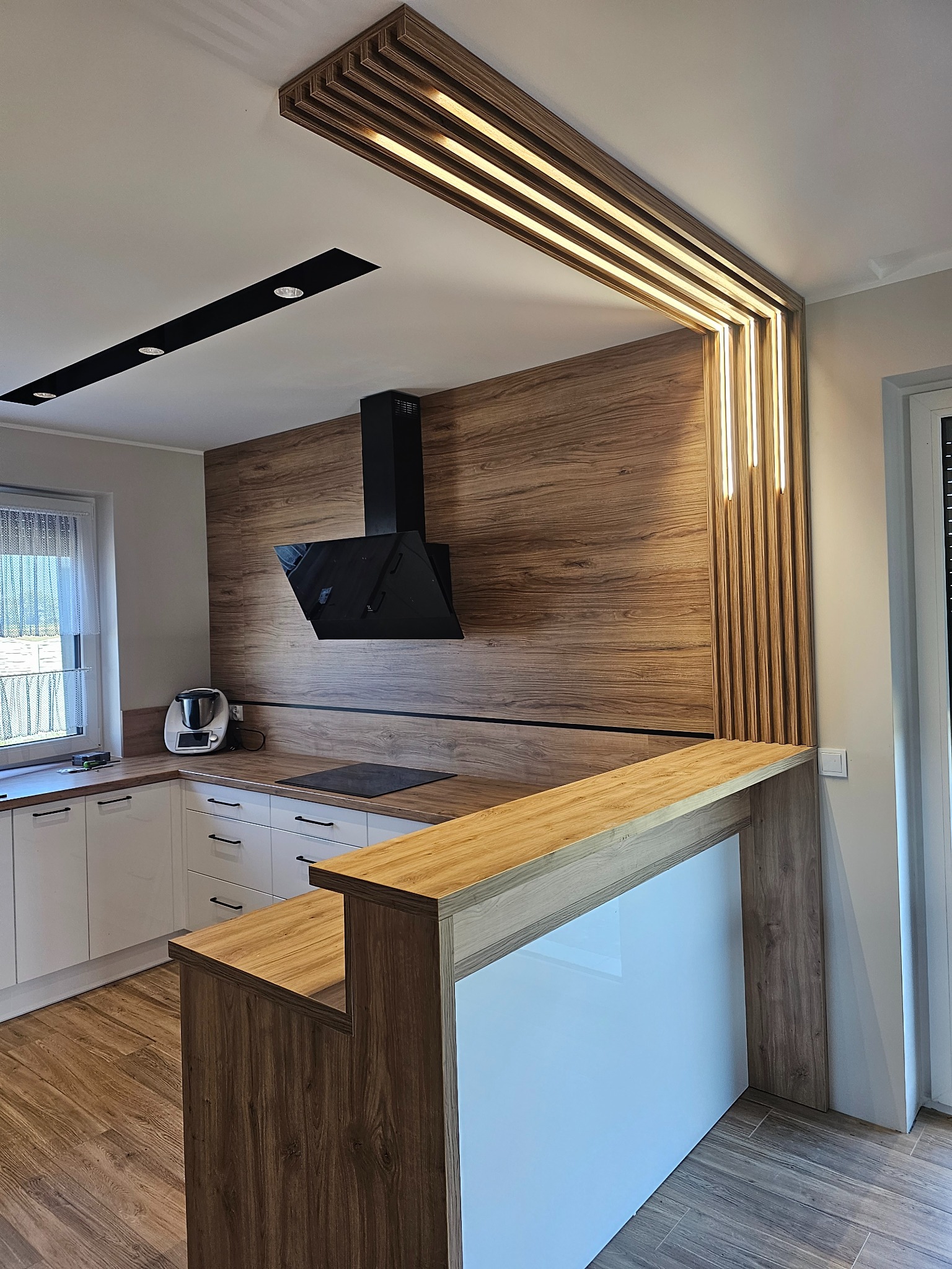 Nowoczesna kuchnia z drewnianymi akcentami: okładzina ściany, blat kuchenny, dekoracyjny element sufitu z oświetleniem LED, białe szafki i czarny okap.