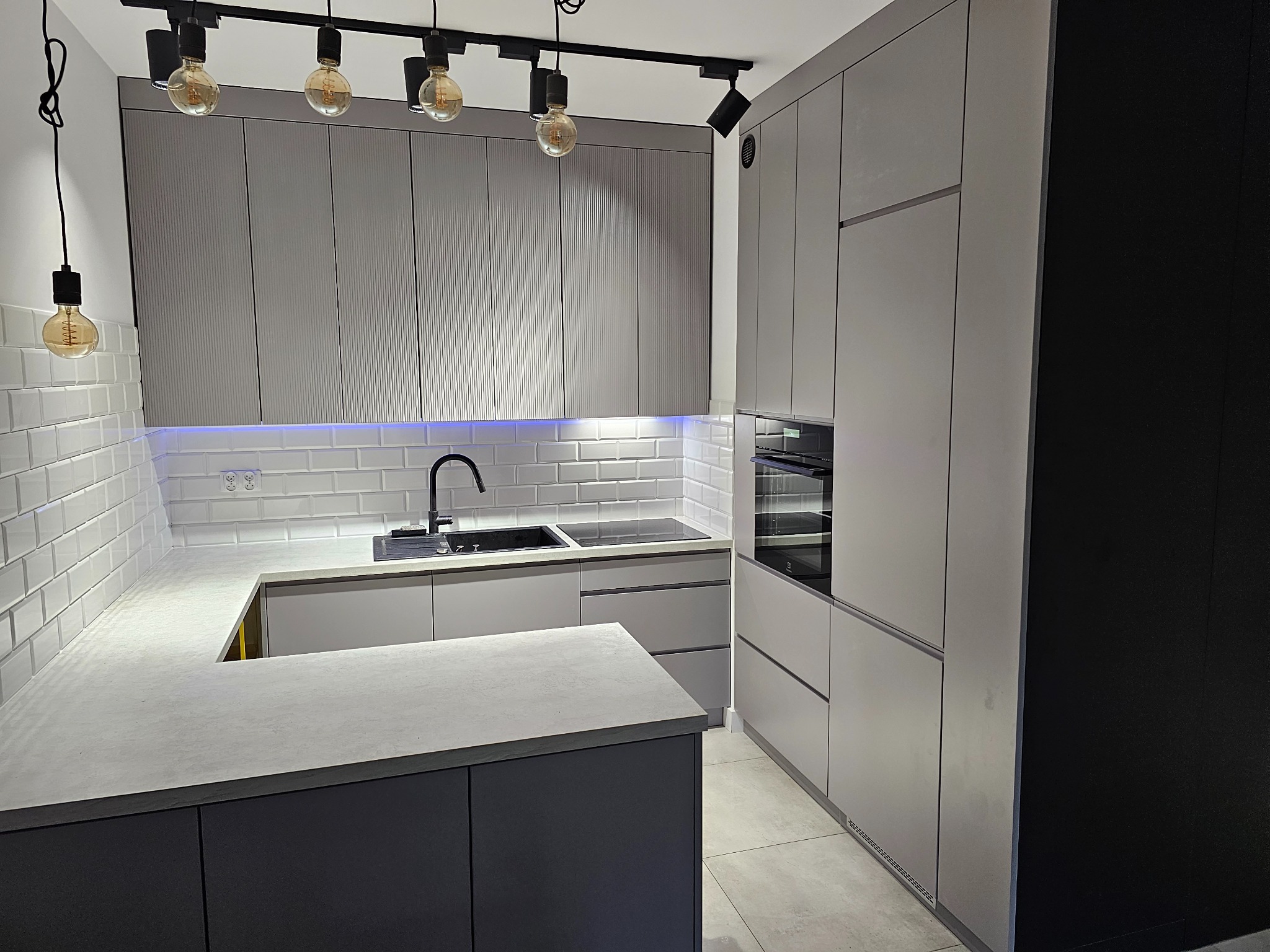 Nowoczesna kuchnia z szarymi frontami, białymi płytkami metra, czarną armaturą i oświetleniem punktowym z żarówkami Edisona.