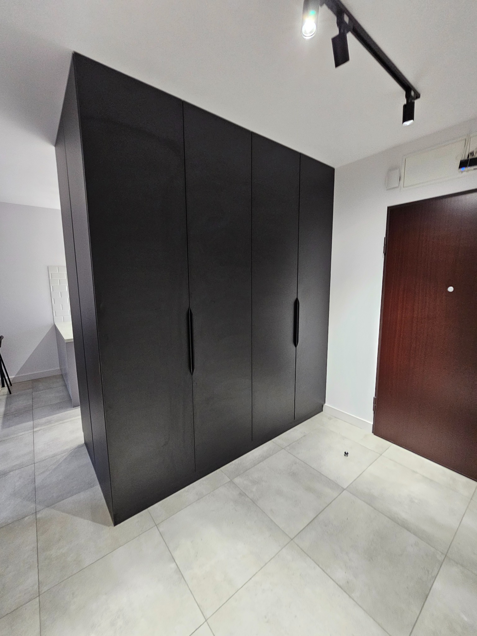 Czarna, matowa zabudowa szafy wnękowej z długimi, pionowymi uchwytami, dopasowana do wysokości pomieszczenia, na tle jasnej podłogi z płytek i białych ścian.