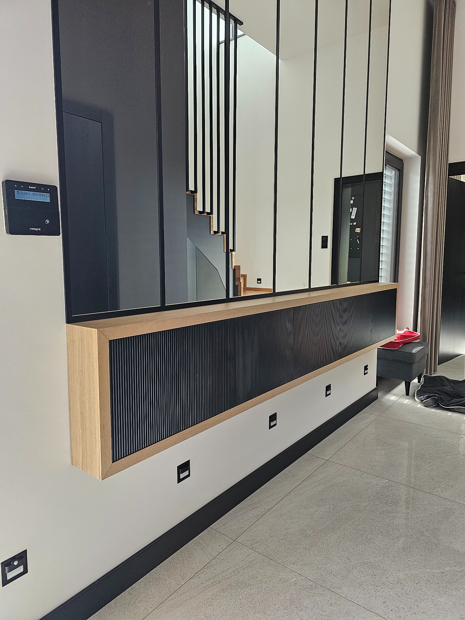 Nowoczesna zabudowa ściany z lamelami w kolorze czarnym, obudowana jasnym drewnem, z przeszklonym panelem górnym, odbijającym schody i fragment wnętrza.