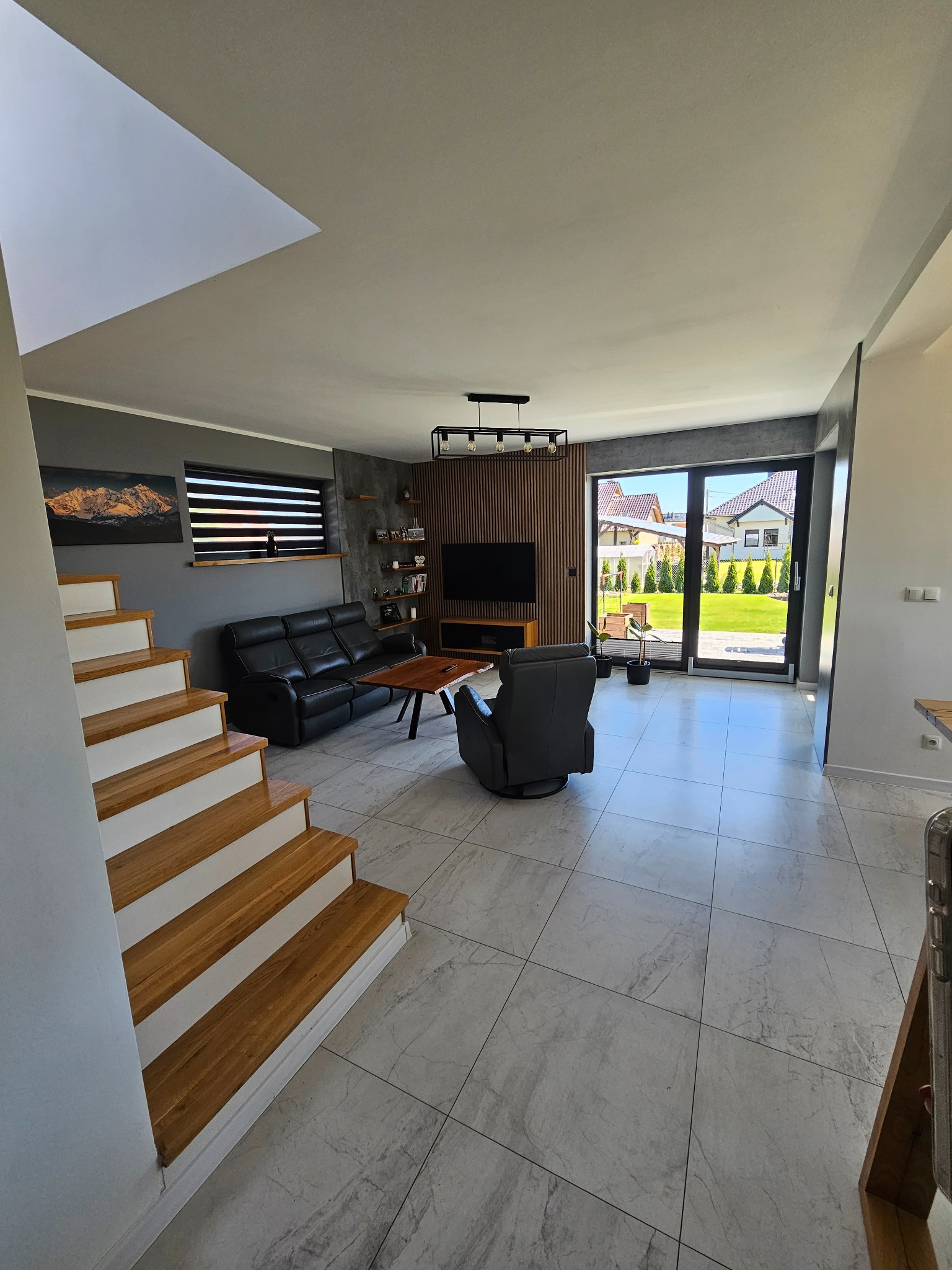 Nowoczesny salon z drewnianymi akcentami: schody z drewnianymi stopniami, ściana z drewnianymi panelami, czarna skórzana sofa i fotel, marmurowe płytki podłogowe oraz widok na zielony ogród przez...