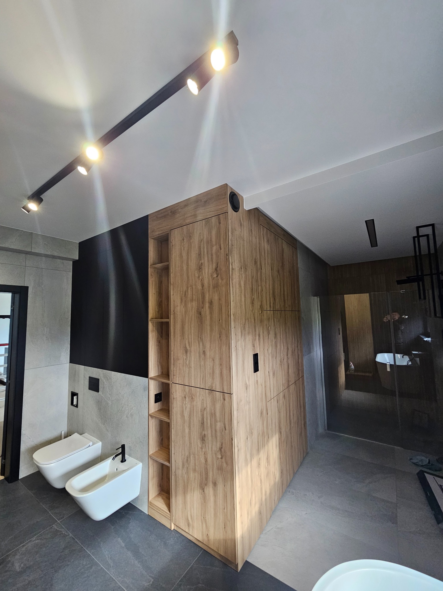 Nowoczesna łazienka z drewnianą szafą wnękową, podwieszaną toaletą i bidetem, oświetlenie szynowe punktowe.