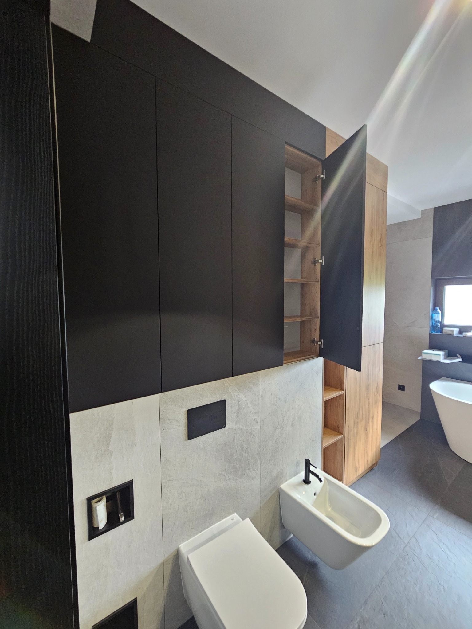 Nowoczesna łazienka z czarnymi szafkami wiszącymi, otwartą szafką z drewnianym fornirem, białą toaletą i bidetem na tle szarych płytek.