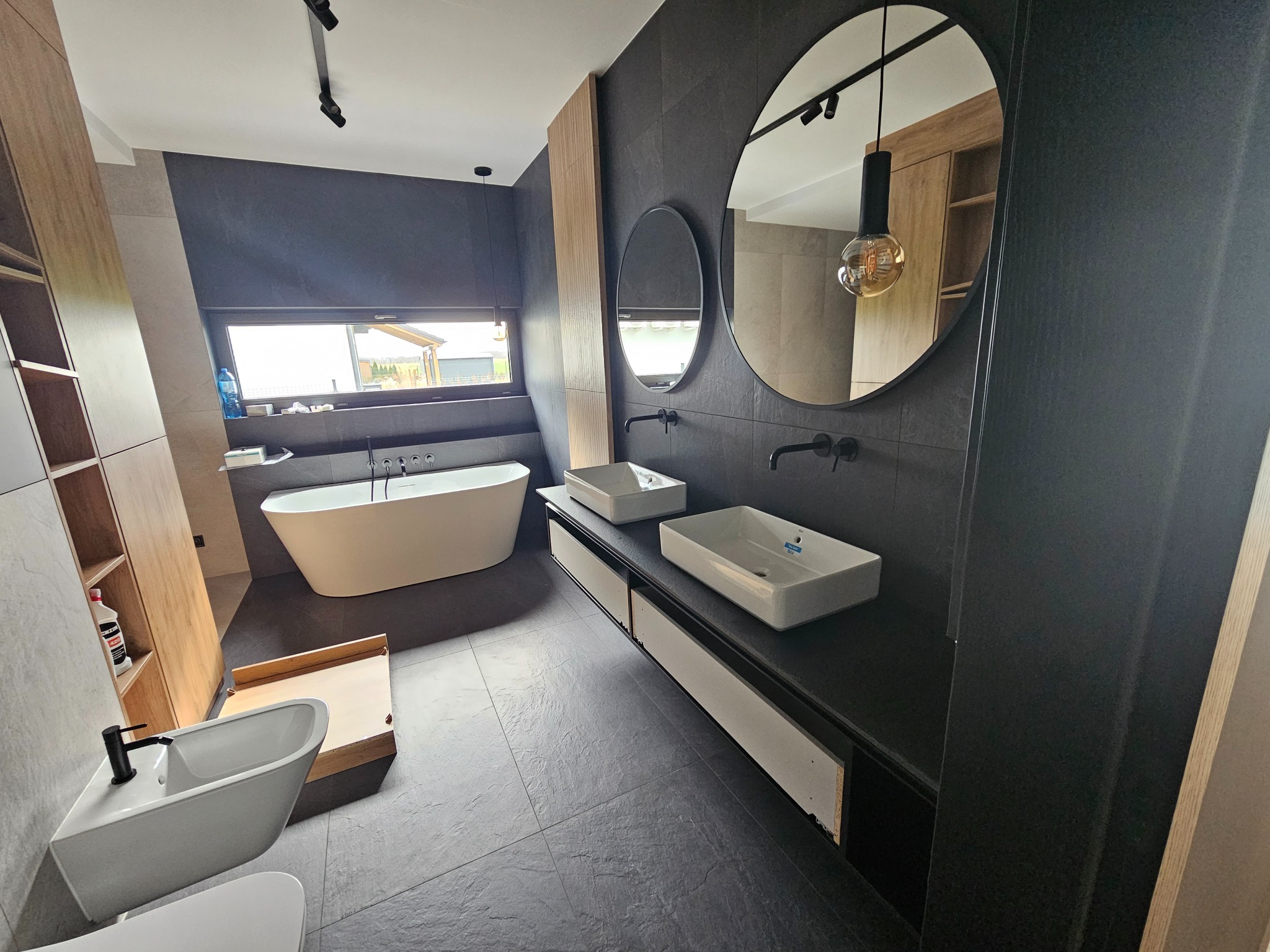 Nowoczesna łazienka z czarnymi płytkami, dwiema umywalkami nablatowymi, okrągłymi lustrami, wolnostojącą wanną przy oknie i drewnianą zabudową szaf.