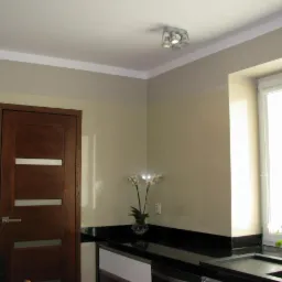 Nowoczesna łazienka z drewnianymi drzwiami z czterema poziomymi przeszkleniami, czarnym blatem, beżowymi płytkami i białym oknem z roletą, oświetlona lampą sufitową z czterema kloszami.