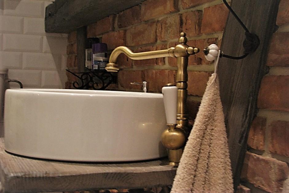 Okrągła, biała umywalka nablatowa z mosiężną baterią w stylu retro, na drewnianym blacie, ceglana ściana w tle, dekoracyjny wieszak na ręcznik.