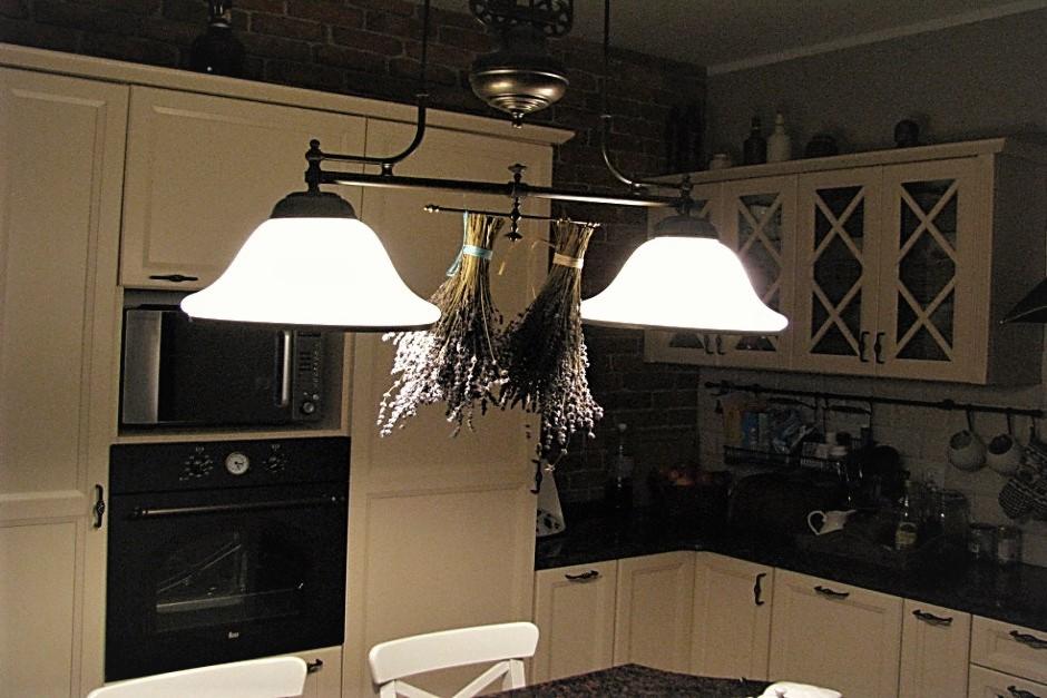 Rustykalna kuchnia z kremowymi szafkami, czarnym blatem i ceglaną ścianą, ozdobiona suszonymi bukietami lawendy wiszącymi pod lampą.