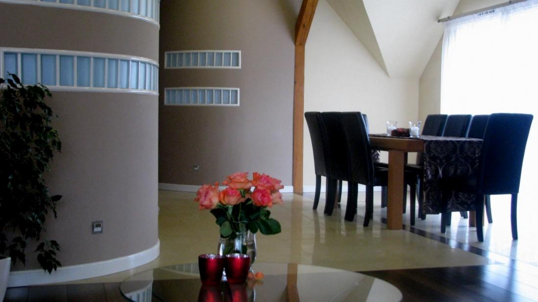 Jasne wnętrze z zaokrąglonym elementem architektonicznym, różowe róże w wazonie na szklanym stoliku, stół z czarnymi krzesłami w tle, drewniany element konstrukcyjny przy ścianie.