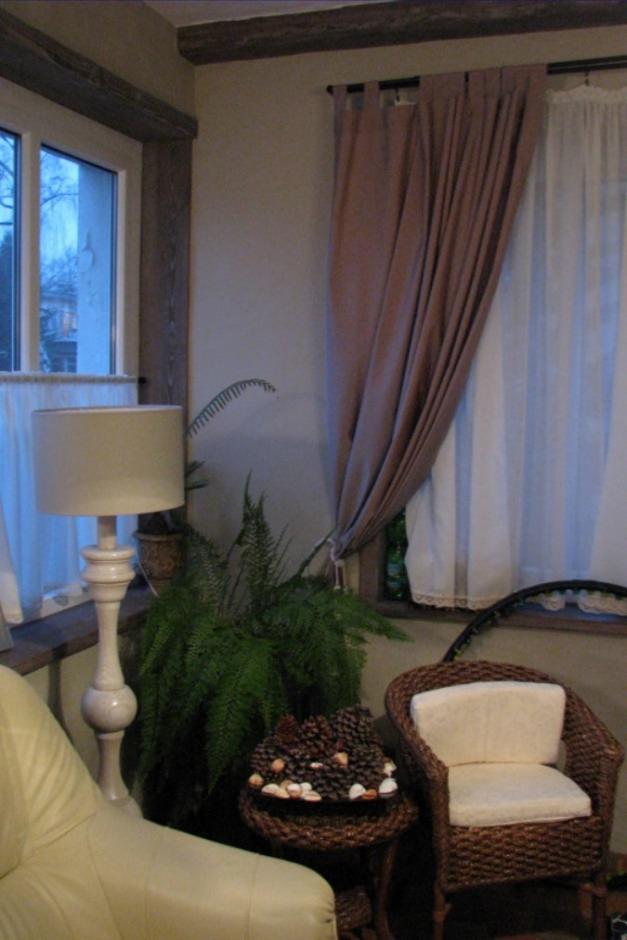 Przytulny kącik z wiklinowym fotelem, stolikiem z szyszkami i muszelkami, wysoką lampą oraz paprotką, doświetlony światłem z okna z beżowymi i białymi zasłonami.