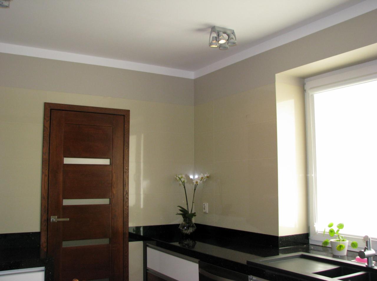 Nowoczesna łazienka z drewnianymi drzwiami z czterema poziomymi przeszkleniami, czarnym blatem, beżowymi płytkami i białym oknem z roletą, oświetlona lampą sufitową z czterema kloszami.