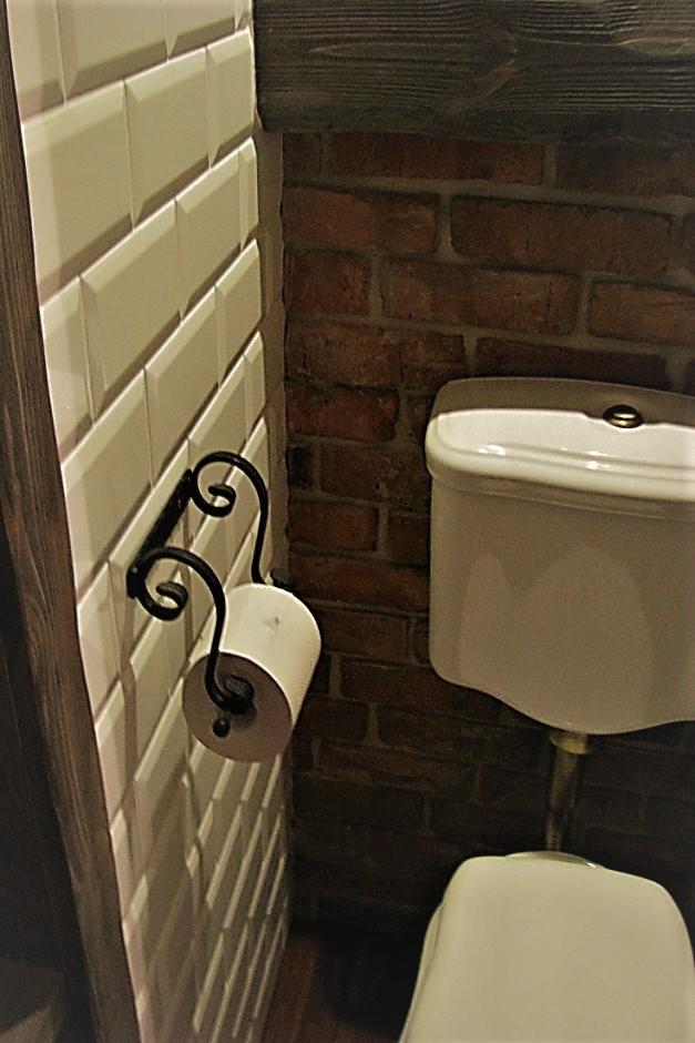 Toaleta w stylu rustykalnym: biała ceramika, ceglana ściana i dekoracyjny uchwyt na papier toaletowy z kutego żelaza na ścianie wyłożonej białymi, podłużnymi płytkami.