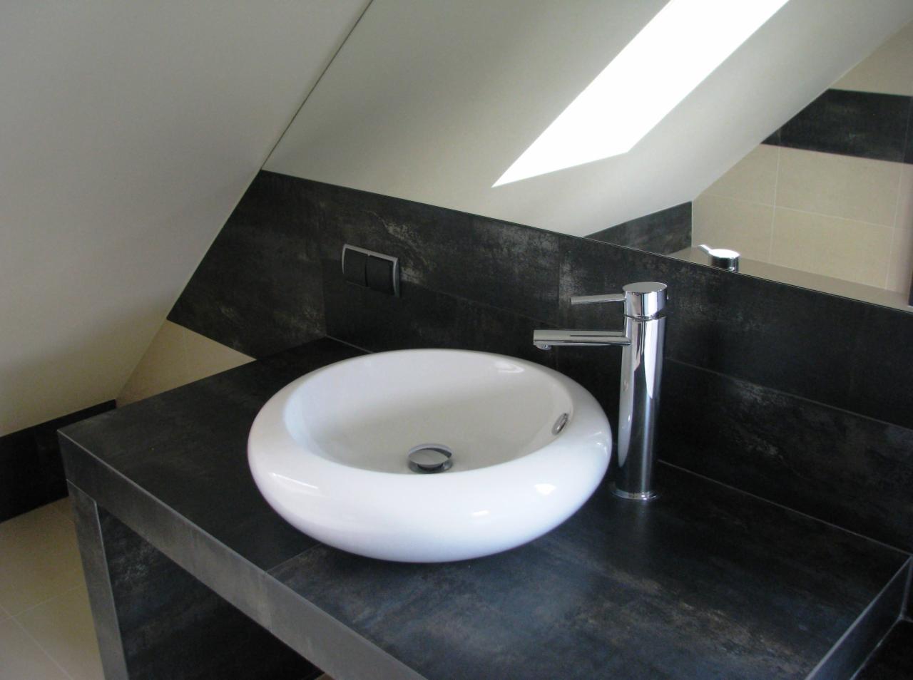 Biała, okrągła umywalka nablatowa z chromowaną baterią na blacie z ciemnego kamienia w łazience na poddaszu, widoczne odbicie w lustrze.