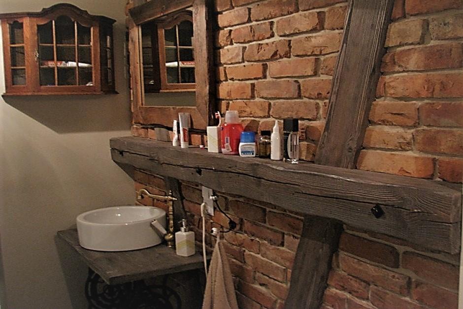 Rustykalna łazienka z umywalką nablatową i półką z surowego drewna na tle ceglanej ściany, uzupełniona o lustro w drewnianej ramie i wiszącą szafkę.