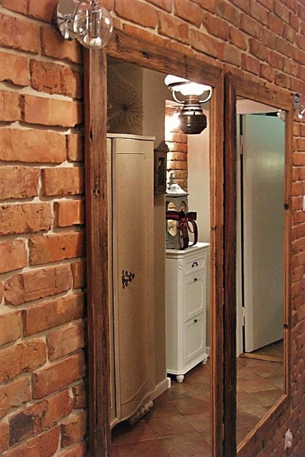 Wnętrze z ceglaną ścianą i lustrem w drewnianej ramie, oświetlone lampą wiszącą, z widoczną szafką i komodą w stylu vintage.