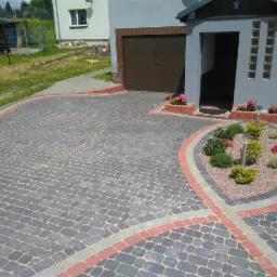 Ułożona kostka brukowa w odcieniach szarości z dekoracyjnymi obramowaniami w kolorze czerwonym i szarym, tworząca wzór na podjeździe przed domem z niewielkim ogródkiem z krzewami i lampami solarnymi.