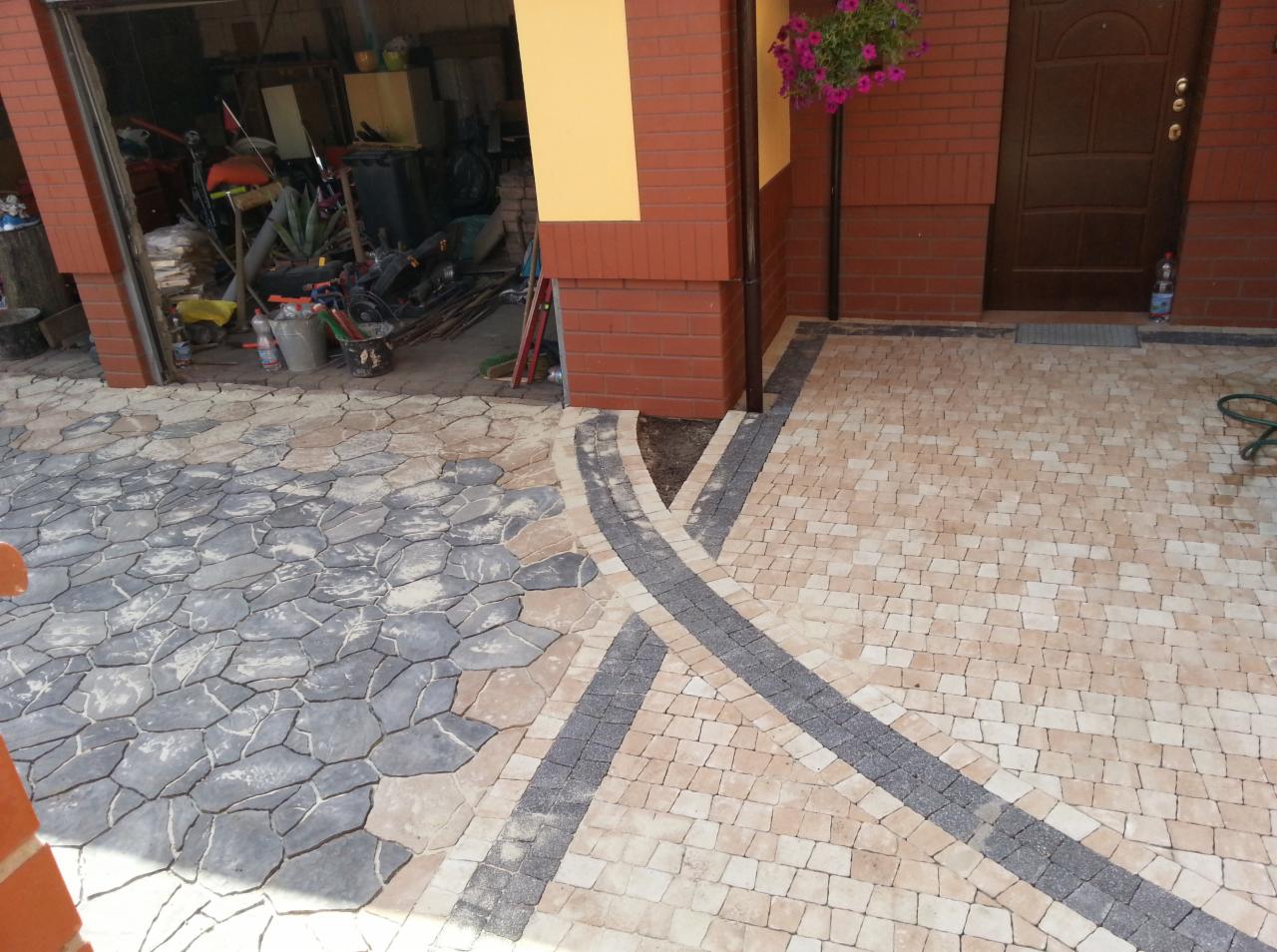 Ułożona nawierzchnia z kostki brukowej w trzech kolorach, tworząca wzór przed wejściem do domu z otwartym garażem, w tle widoczne narzędzia i materiały budowlane.
