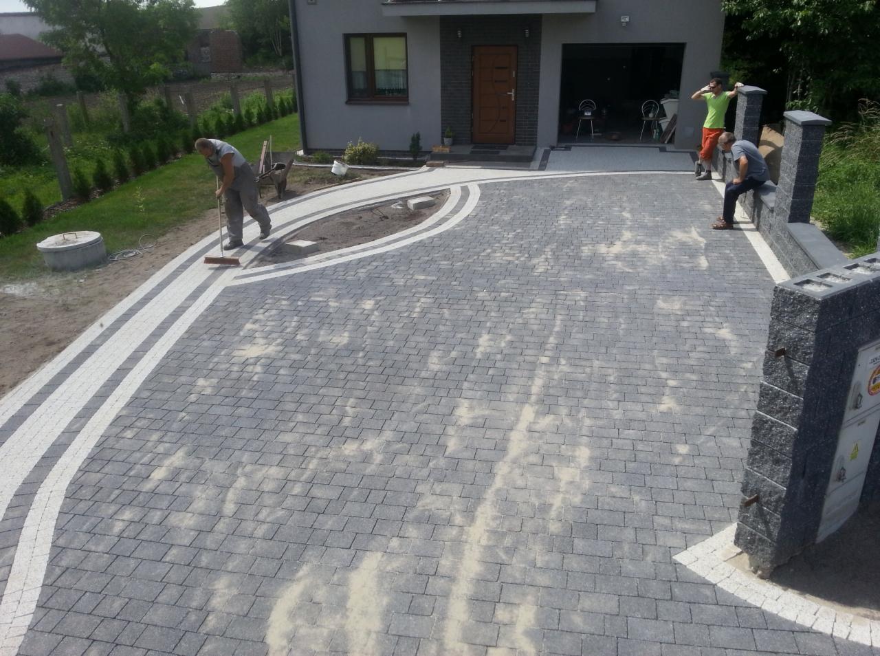 Pracownik zamiata piasek z nowo ułożonej, szarej kostki brukowej z białymi obrzeżami na podjeździe przed domem z garażem; w tle dwóch mężczyzn obserwuje prace.