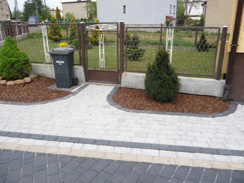 Ułożona jasna kostka brukowa z obramowaniem z ciemnej kostki, rabaty z korą i iglakami, metalowa brama i ogrodzenie, widoczny kosz na śmieci.