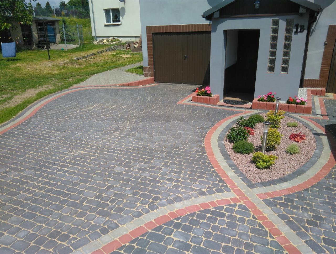 Ułożona kostka brukowa w odcieniach szarości z dekoracyjnymi obramowaniami w kolorze czerwonym i szarym, tworząca wzór na podjeździe przed domem z niewielkim ogródkiem z krzewami i lampami solarnymi.
