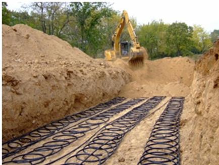 Żółta koparka wykonująca wykop pod instalację rur spiralnych w ziemi, widok z góry na ułożone rury w rowie.