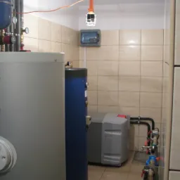 Kompletna kotłownia: szary zasobnik Sofath z tarczą, niebieski bojler i szary piec w kafelkowanym pomieszczeniu. Widoczne rury i zawory.