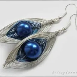 Para ręcznie robionych kolczyków z niebieskimi perłami w srebrnych, drucianych oprawach, ułożonych na jasnym tle.