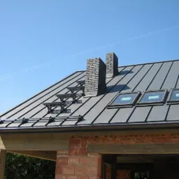 Szary dach z blachy trapezowej z trzema oknami dachowymi i ceglanym kominem w słoneczny dzień, widoczne schodki dachowe i rynny.