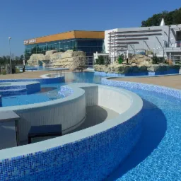 Kompleks basenowy z mozaiką w odcieniach błękitu i bieli, fontanną i dekoracyjnymi elementami skalnymi na tle nowoczesnego budynku z przeszkloną fasadą.