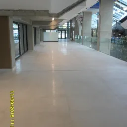 Jasna, wyłożona płytkami posadzka przemysłowa w długim, przestronnym korytarzu biurowym z przeszklonymi ścianami i widokiem na zieleń. Widoczna data i godzina na dole zdjęcia.