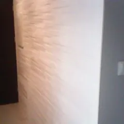 Biała ściana z dekoracyjną fakturą imitującą rozchodzące się promienie, widoczny fragment szarej ściany z włącznikiem i ciemne drzwi w tle. Minimalistyczny design.