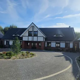 Nowoczesny dom z elementami muru pruskiego, czarną dachówką i brukowanym podjazdem w kształcie ronda z nasadzeniami.