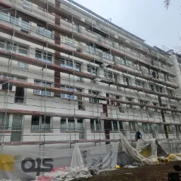 Wielopiętrowy budynek w trakcie ocieplania elewacji, otoczony rusztowaniami, z widocznymi pracownikami i zabezpieczeniami w postaci folii ochronnej z logo firmy Sto, przy pochmurnej pogodzie.