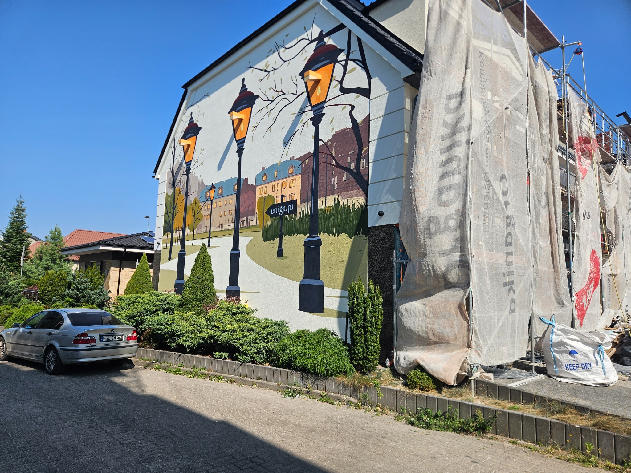 Ściana budynku z namalowanym muralem przedstawiającym uliczne latarnie i budynki, częściowo zasłonięta rusztowaniem i plandeką podczas prac remontowych, obok zaparkowany srebrny samochód.