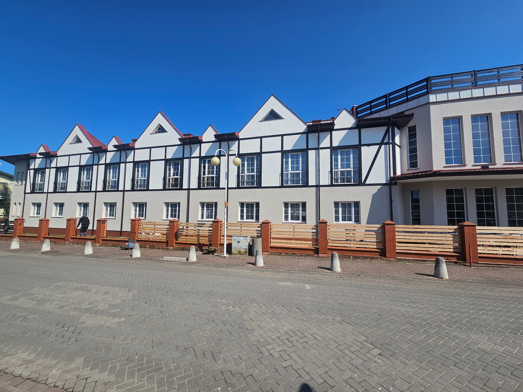 Dwukondygnacyjny budynek z elementami szachulcowymi, czerwonym dachem i jasnoszarą elewacją, otoczony niskim płotem z ceglanymi słupkami, widziany w słoneczny dzień.