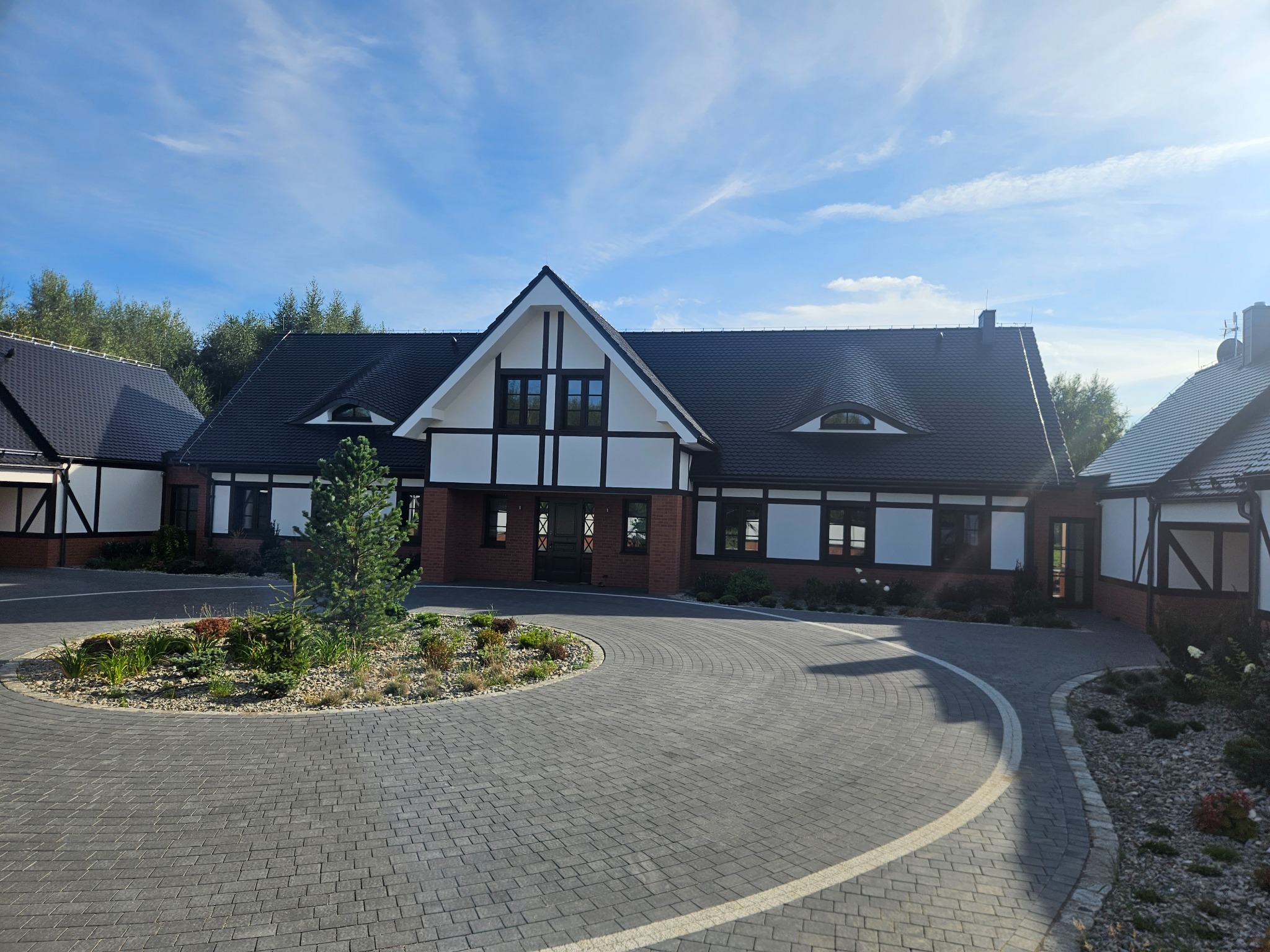 Nowoczesny dom z elementami muru pruskiego, czarną dachówką i brukowanym podjazdem w kształcie ronda z nasadzeniami.