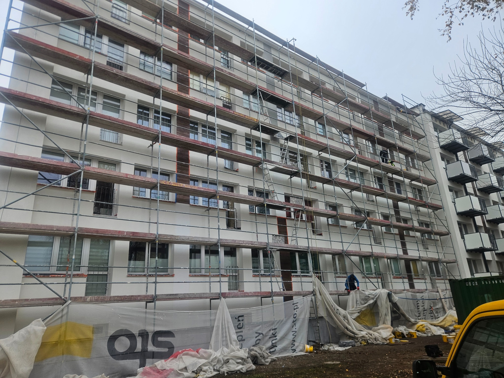 Wielopiętrowy budynek w trakcie ocieplania elewacji, otoczony rusztowaniami, z widocznymi pracownikami i zabezpieczeniami w postaci folii ochronnej z logo firmy Sto, przy pochmurnej pogodzie.