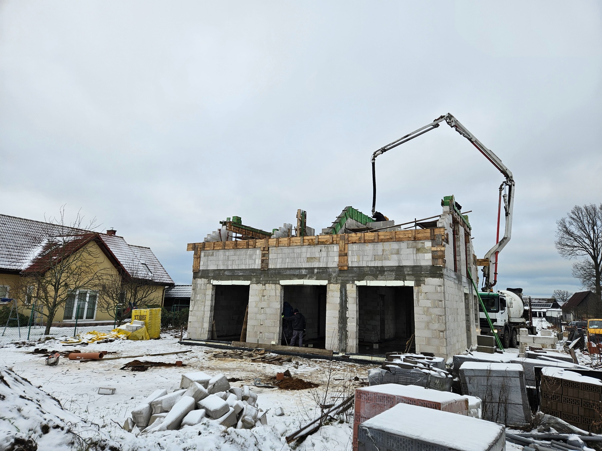 Surowy stan domu z betonu komórkowego w zimowej scenerii, widoczny betonowóz z pompą podającą zaprawę, palety z bloczkami Ytong przykryte śniegiem, obok stoi żółty dom.