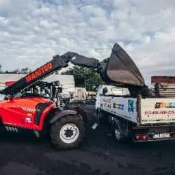 Czerwona ładowarka teleskopowa Manitou nabiera węgiel do skrzyni samochodu ciężarowego z reklamą składu opału.