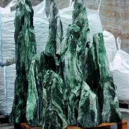 Zielone, strzeliste kamienie dekoracyjne z wyraźnym wzorem żyłkowania, ułożone na drewnianej palecie, w tle białe worki typu big bag.