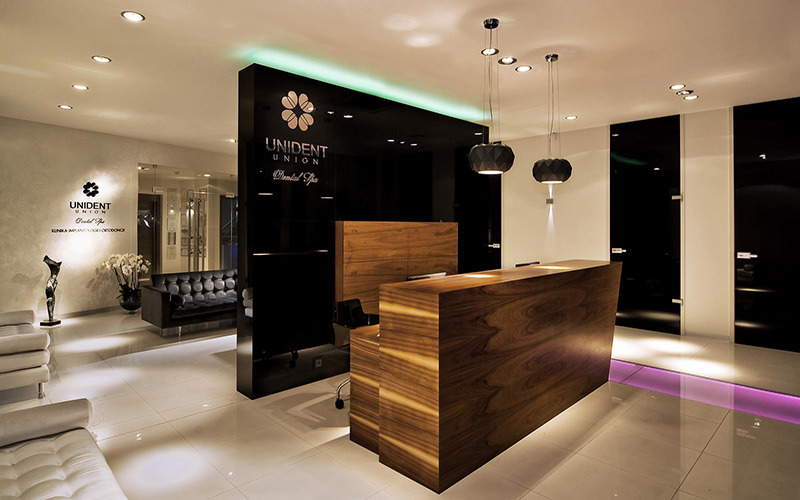 Nowoczesne wnętrze recepcji kliniki dentystycznej Unident Union z drewnianą ladą, czarnymi ścianami i designerskimi lampami wiszącymi.